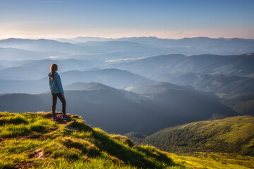 Girl on mountain peak with green grass looking at beautiful mountain valley in fog at sunset in summer. Landscape with sporty young woman, foggy hills, forest, sky. Travel and tourism. Hiking