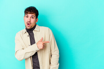 Wall Mural - Young caucasian man isolated on blue background smiling and pointing aside, showing something at blank space.