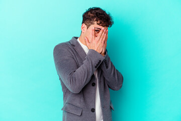 Wall Mural - Young caucasian man isolated on blue background blink through fingers frightened and nervous.