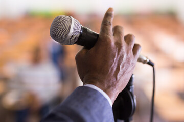 Closeup of microphone in hand of African American man on blurred background of conference room. Concept of speaking during public event