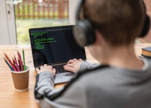 Young Boy Hacking And Coding System On Computer Laptop With Green Program Code Text And Selective Focus On Keyboard Wearing Headphones Sat At Desk
