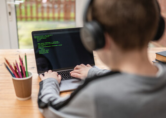 Young boy hacking and coding system on computer laptop with green program code text and selective focus on keyboard wearing headphones sat at desk