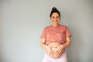 Wall Mural -  Smiling pregnant woman holding her belly and making a heart shape. Looking at camera.