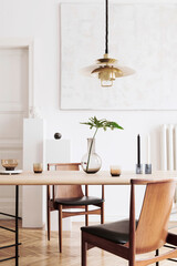 Stylish and modern dining room interior with design sharing table, chairs, gold pendant lamp, abstract paintings and elegant accessories. Tropical leafs in vase. Eclectic home decor.