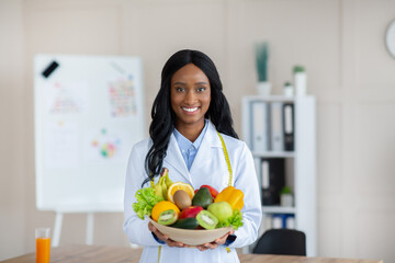 Portrait of happy black dietitian in lab coat holding bowl of fresh fruits and vegetables, smiling at camera at clinic