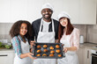 © sofiko14 - Happy smiling parents, African man, Caucasian woman and their mixed race teen child, holding baking tray with fresh tasty pastries. Cook at home and family bakery concept