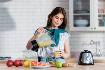 young woman pouring smoothie from blender near fresh fruits in kitchen