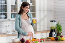 Happy Pregnant Woman Touching Tummy While Holding Fresh Juice In Kitchen