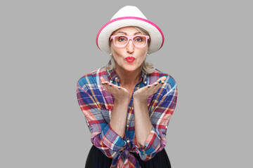 Portrait of funny modern stylish mature woman in casual style with hat and eyeglasses standing sending kiss and looking at camera with romantic expression. studio shot isolated on gray background.