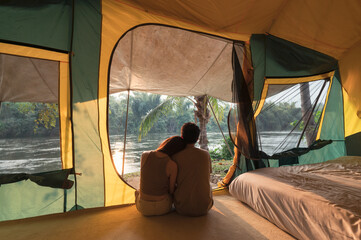 Wall Mural - Young asian couple embracing and relaxing inside a tent on campsite in natural park
