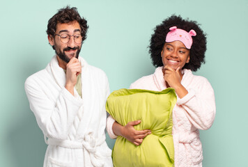 Wall Mural - multiracial couple of friends smiling with a happy, confident expression with hand on chin, wondering and looking to the side. pajamas and home concept