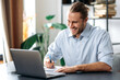 © Kateryna - Handsome caucasian guy freelancer uses laptop. Smiling attractive modern man in stylish casual wear sits at the desk in a creative or home office, browsing internet, replies to email, distant learners