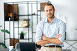 Handsome confident attractive caucasian stylishly dressed guy, designer, freelancer or employee, standing near his desk, with arms crossed, looking at the camera and smiling friendly