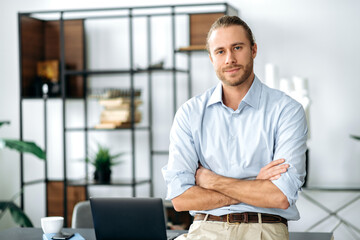 Handsome confident attractive caucasian stylishly dressed guy, designer, freelancer or employee, standing near his desk, with arms crossed, looking at the camera and smiling friendly