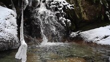 Water Clattering Down At Black Rocks On A Small Waterfall In A Winter Scene At A Creek With White Snow At The Sides And Ice Dangling Above Water. Slowmo Pedestal Shot