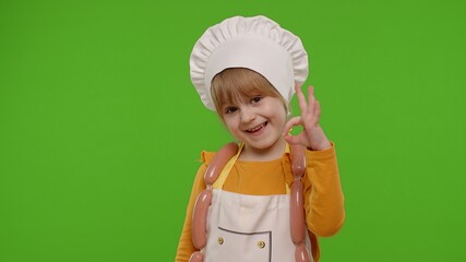 Wall Mural - Charming child girl dressed in apron and hat like chef cook showing approval ok sign, invites you to come on chroma key background. Nutrition, family cooking school, children education, restaurant