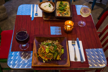 Wall Mural - vegan cuisine with drinks on a table set for two. These healthy options are Thai Green Curry with white rice and Pad Thai. Glasses of white wine and beer are also on the candlelit table