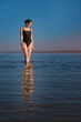 © Roman - Young beautiful slim woman wearing a black swimsuit stands in the sea and watching sunset. Copy space vertical