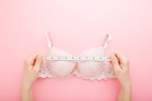 Young Adult Woman Hands Holding White Measure Tape And Measuring Bra On Light Pink Table Background. Pastel Color. Closeup. Point Of View Shot. Choosing Right Size. Top Down View.