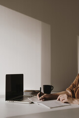 Wall Mural - Women's hand write in a blank sheet notebook. Minimalist home office desk workspace. Laptop on table with coffee cup, paper sheet, stationery against white wall. Work, business background.