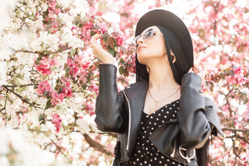 Wall Mural - Young woman in flowered park rests in nature. Fashionable girl in elegant hat in trendy sunglasses in black outfit and pink spring flowers outdoors. Luxurious lady sniffs amazing flowering tree.