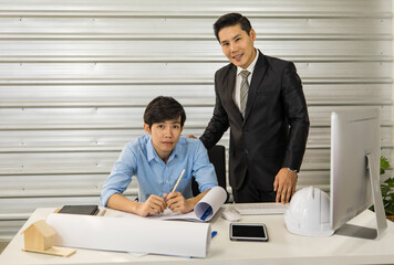 Asian senior good-looking boss in suit sitting at desk and working with junior employee in office with roll of paper. Businessmen teamwork and good colleagues concept