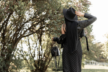 Wall Mural - Back view on fashionable woman with healthy long chic hair in elegant black hat in beautiful dress in vintage jacket with leather handbag in park. Trendy girl in fashion black spring clothes on walk.