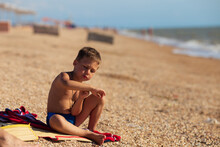 A Boy Sits On A Red Towel Of A Sandy Beach Near The Sea And Looks At His Hand
