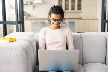 Wall Mural - Positive intelligent African American teenage girl student in glasses sitting on the couch with laptop on the lap, studying from home, typing email, writing article, working on project, watching video