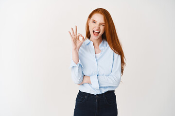 Wall Mural - Cheerful redhead female manager showing OK sign and winking confident, give assurance, saying yes, praising good deal, standing over white background