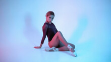 Young Ballerina In Bodysuit And Pointe Sitting On Blue And Pink Background