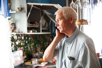 senior grandfather talking with phone near window at home. elderly man sad. grandfather chatting with relatives or grandchildren. Soft focus