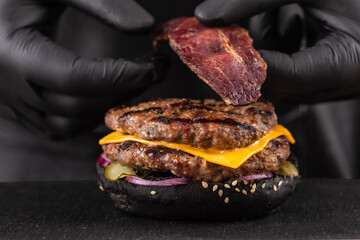 Wall Mural - Close-up, making grilled burger. Chef hands in black gloves are puts slice of bacon in burger with double beef cutlet.