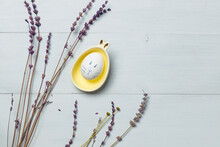 An Easter Egg With A Painted Rabbit Face, In A Yellow Bowl With Ears. On A Wooden Background With Lavender Flowers