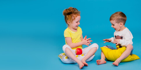 Positive cute toddler and redhead curly girl on the pillows are playing with apple on the blue background.Summer vacation and holidays concept,travel with children.Copy space for mock up
