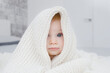 Cute little caucasian amazed baby playing hiding under white knitted blanket at home in light interior. Kid games, having fun indoors and winter cold idea