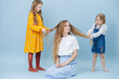 © zzzdim - Two little girls playing with their mother's hair over blue background. One making braid, while second one making hair mustache and laughing. Happy mom looks at camera.