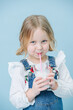 © zzzdim - Funny little blonde girl in overalls and fluffy white shirt holding glass of milkshake, making faces, grimacing, grining over blue background