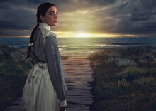 Young Victorian Girl In White Dress And Blue Striped Blouse On The Coast At Sunset.
