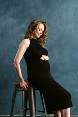 Smiling pregnant brunette young woman with long curly hair, in elegant black midi dress. Fashion style studio photoshoot of pregnancy. Between the second and third trimesters of pregnancy