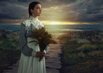 Wall Mural - Young victorian girl holding dried flowers in white dress and blue striped blouse at the coast at sunset.