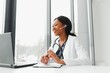 © Serhii - African American woman doctor working at her office online using portable inormation device. Telemedicine services. Primary care consultations, psychotherapy, emergency services.