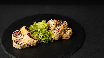 Wall Mural - Custom sushi roll in tempura with nori, fresh salmon, tuna, avocado, masago caviar, drizzled with pineapple sauce with salad pouring as decoration on a black plate on a black table and background.