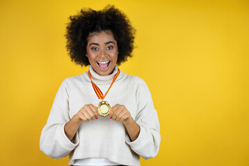 African american woman wearing casual sweater over yellow background holding a medal with a happy face standing and smiling with a confident smile showing teeth