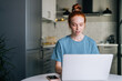 Portrait of pretty redhead young woman working typing on laptop computer sitting at table in kitchen room. Concept of remote working from home office during quarantine from COVID-19.