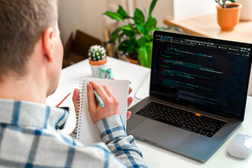 Wall Mural - over his shoulder, a close-up view of a man writing in a notebook, a programmer using a laptop compu