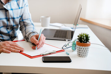  A young man in a shirt in a bright office works at a laptop and makes notes in a notebook, the mental activity of a project manager or an application developer