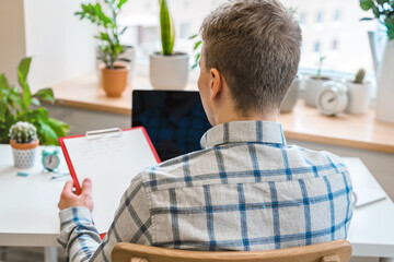 Canvas Print - a young man in a shirt in a bright office works at a laptop and makes notes in a notebook, the menta