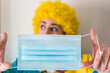 © AliciaFdez - Linda mujer joven sonriendo mientras está vestida con un disfraz de payaso y una peluca de payaso amarilla. Sujeta una mascarilla azul desechable  contra el virus sars-cov-2