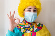 © AliciaFdez - Linda mujer joven sonriendo mientras está vestida con un disfraz de payaso con una mascarilla quirúrgica desechable y una peluca de payaso amarilla. Protección contra el virus sars-cov-2